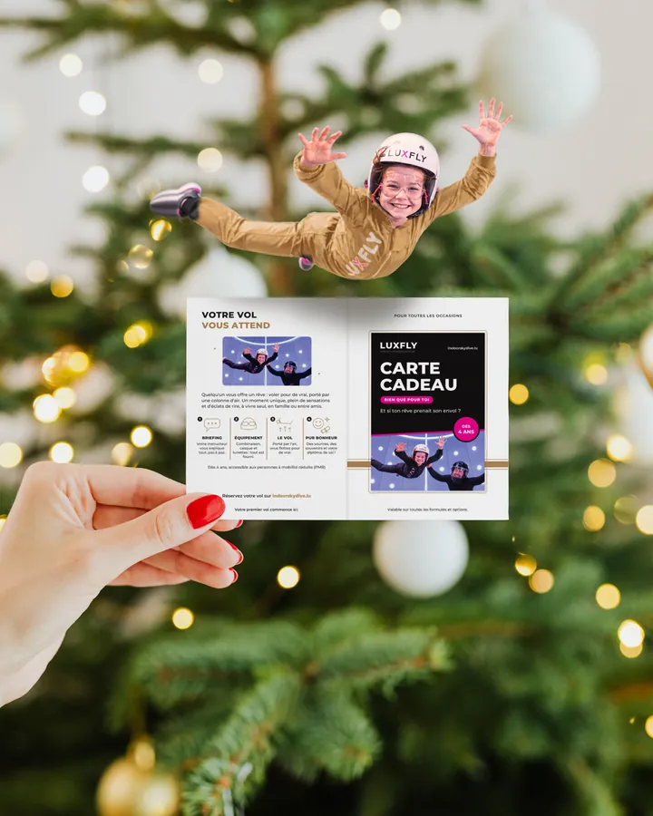 Une petite fille vole au-dessus de la pochette-carte cadeau LUXFLY ouverte, devant un sapin de No&euml;l
